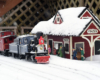 A blue 4-4-0 steam locomotive and its caboose sit at a small wooden depot as snow falls