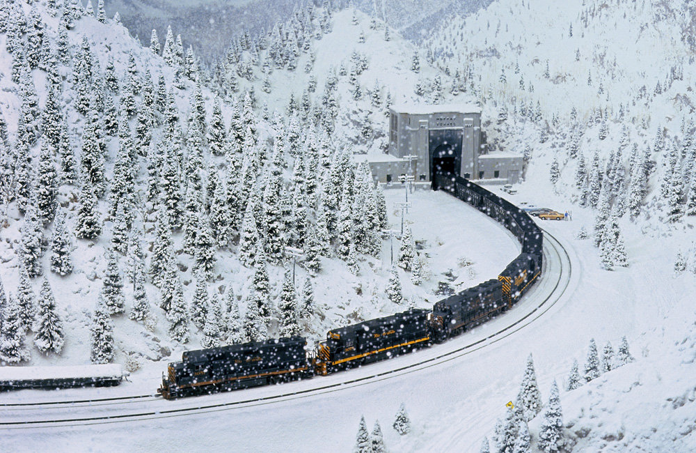 As snowflakes whirl in the sky, a black coal train emerges from a tunnel into a white Rocky Mountain scene
