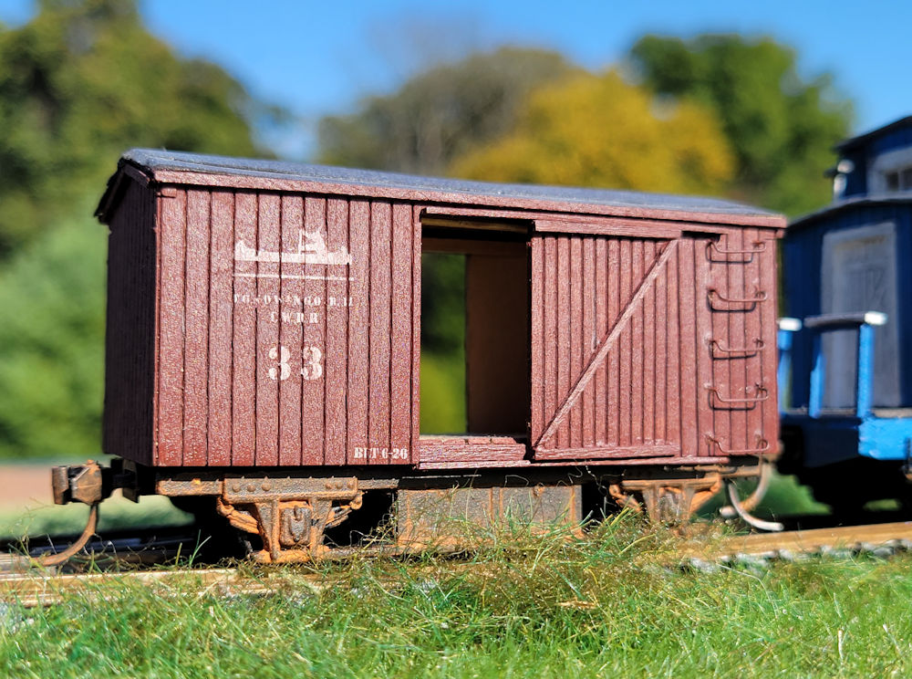 A red model boxcar is shown on model railroad tracks outdoors
