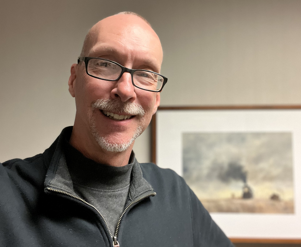 Meet David Popp: Balding middle-aged white male standing in front of a blurry painting of a steam train.