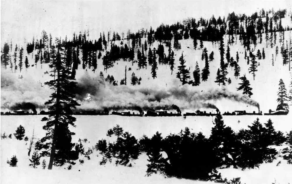 Seven small steam locomotives pushing a wedge snow plow in the mountains