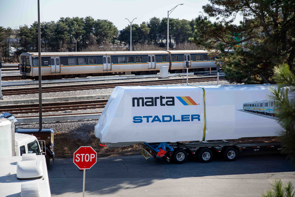 Rapid-transit carbody in wrapper sits on trailer while MARTA train sits in background