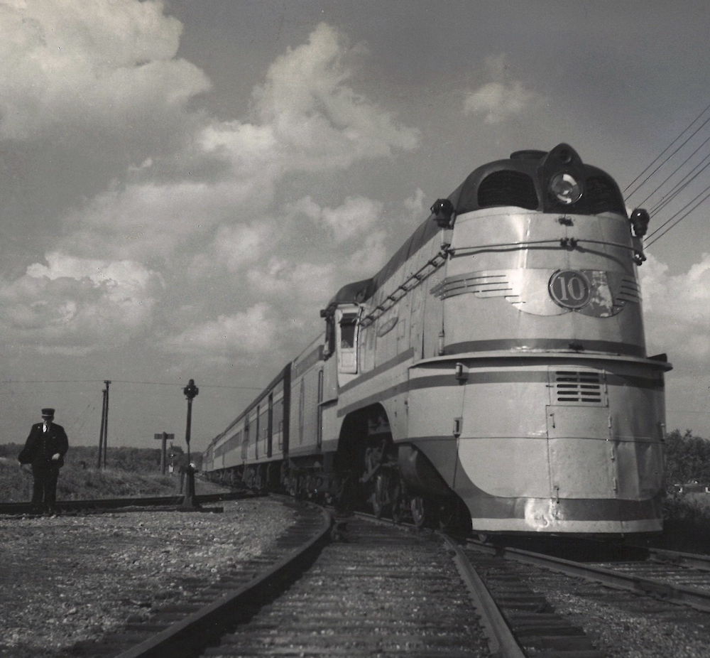 Streamlined steam locomotive backing a passenger train with conductor following.