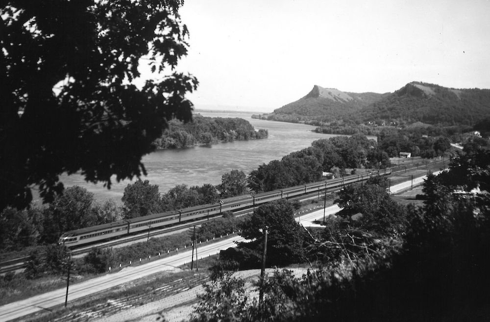 Streamlined passenger train traveling alongside a river in black and white.