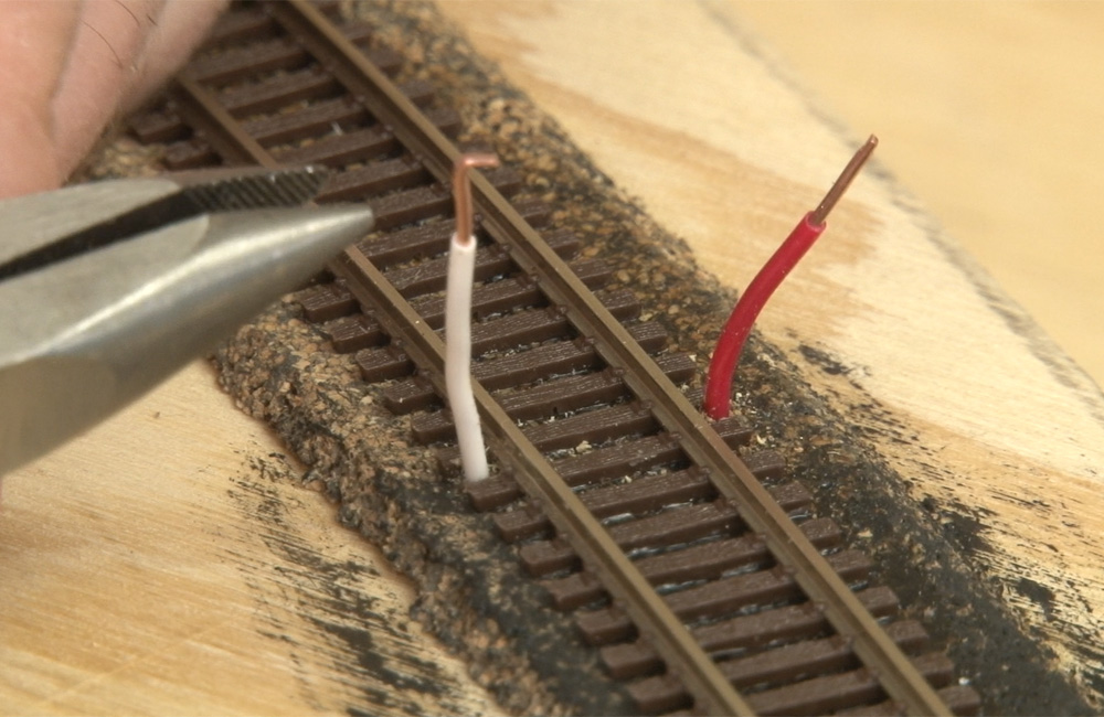 A pair of needle-nose pliers reach toward red and white wires that stick up on either side of a section of N scale flextrack