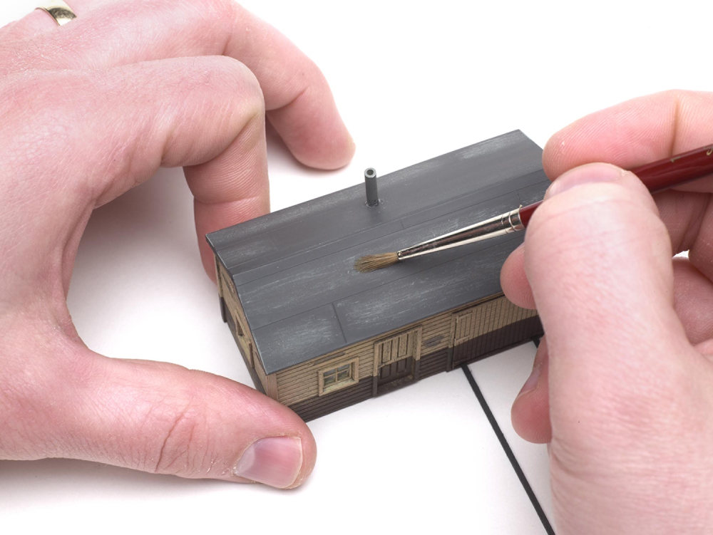 Weathering structures with drybrushing: A pair of hands holding a paintbrush is used on the roof of a structure