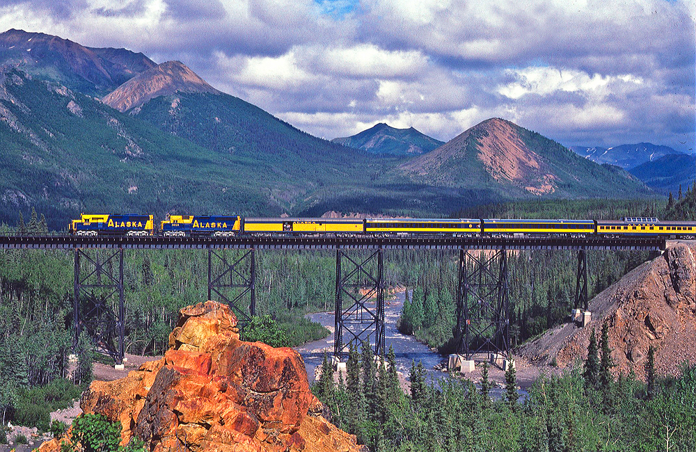 Alaska Railroad passenger train crossing high bridge with mountains in back ground