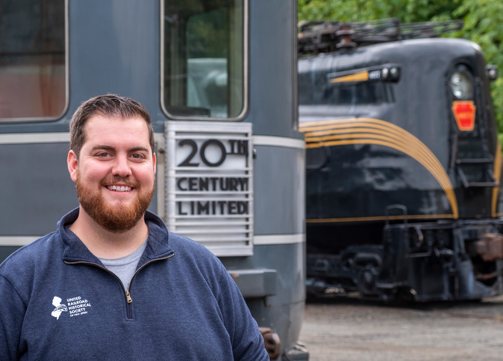 Man standing next to end of observation car with GG1 locomotive in back ground