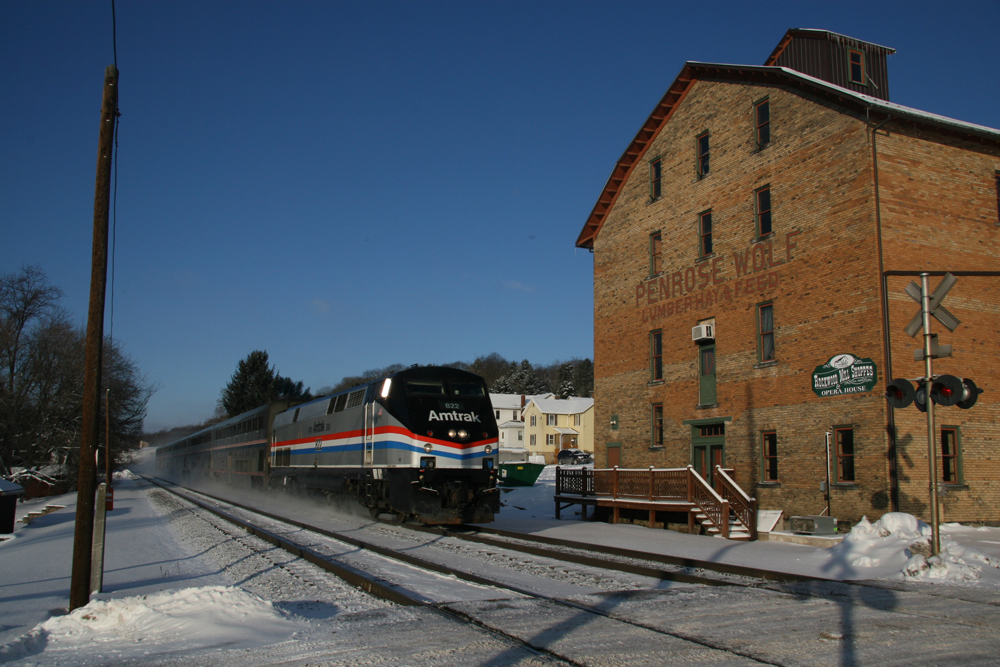 Passenger train passes four-story brick building in snow-covered landscape