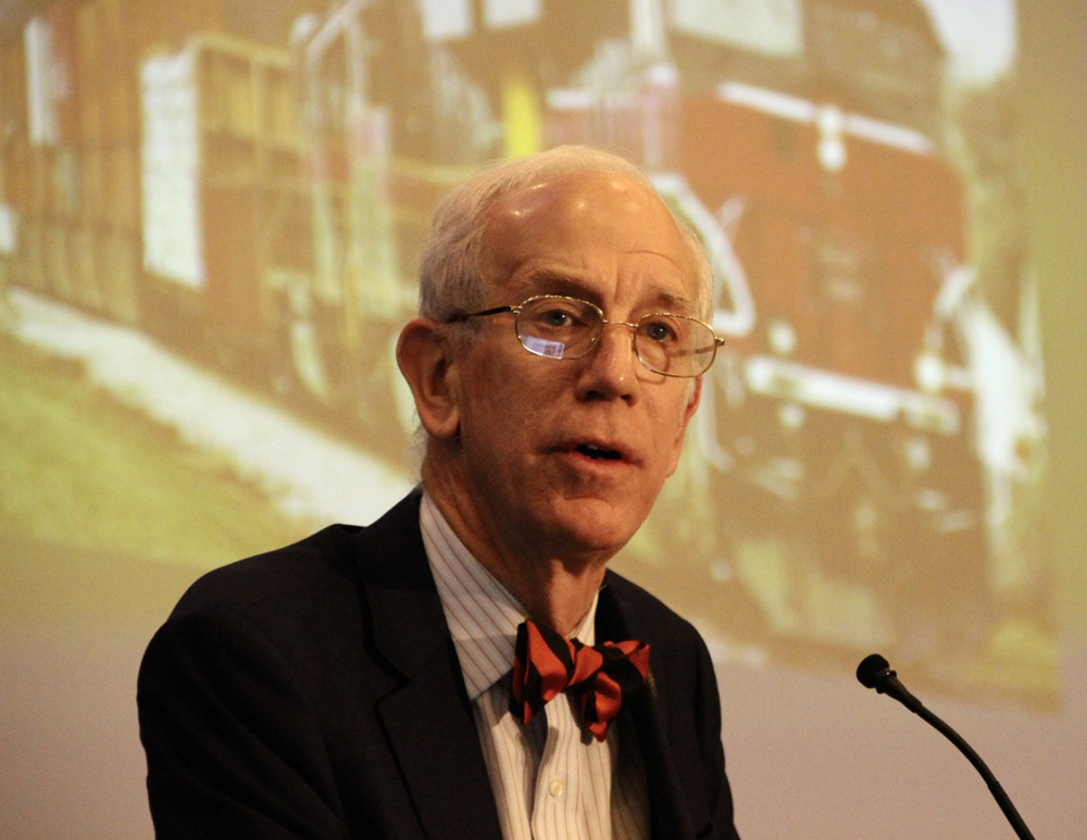 Man with bow tie speaking at microphone with photo of train in background