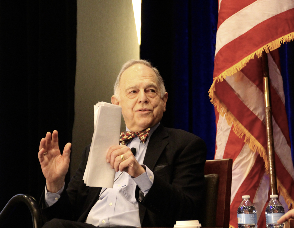 Man holding papers gestures while seated next to American flag