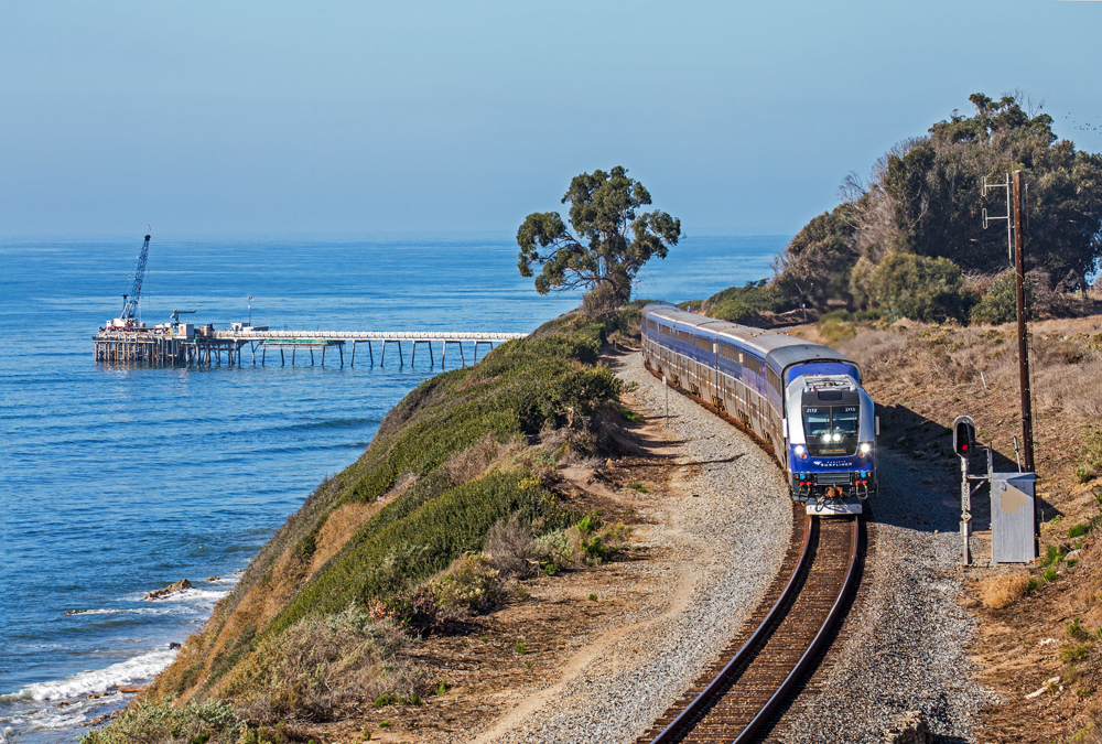 Passenger train running along Pacific coast with pier for oil field in background