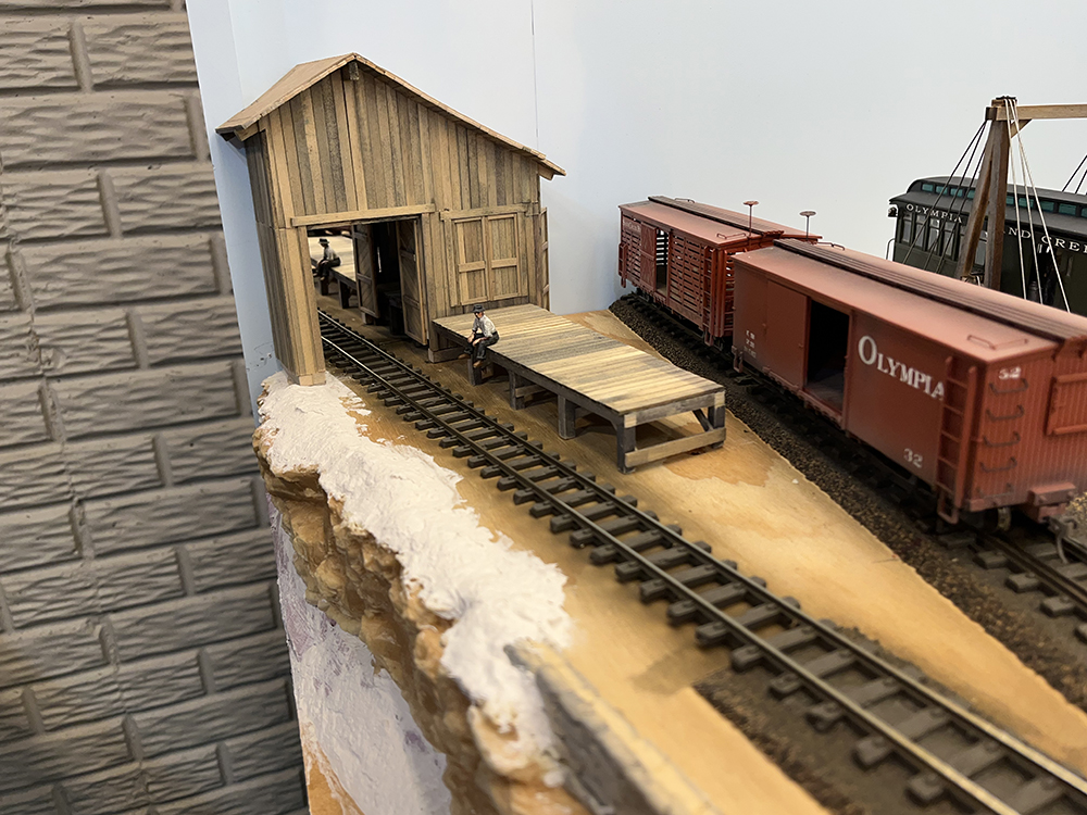 Add depth to structures using mirrors: Brown wood building with long wood loading dock along a stretch of track that appears to run through an open door in the building.