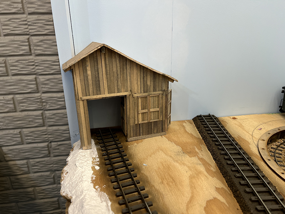 Add depth to structures using mirrors: Brown wood model building against a light blue wall with a brown railroad track entering its open doorway.