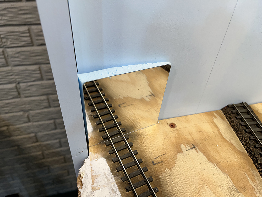 Small mirror supported by blue foam block attached at angle to blue wall and reflecting brown model railroad track.