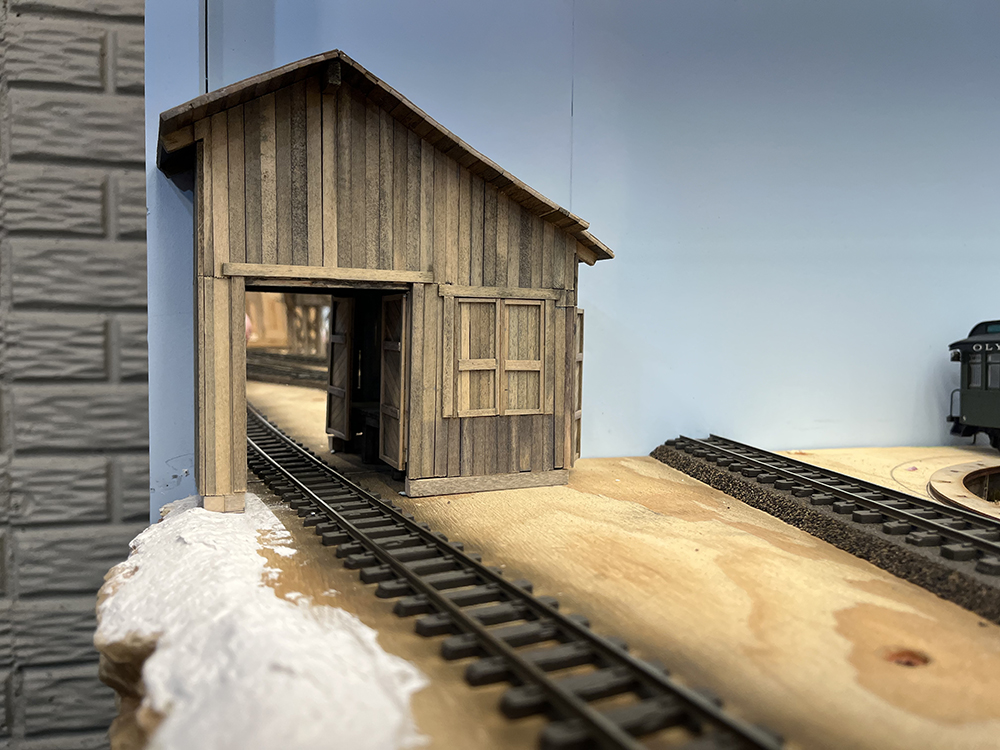 brown model building against blue sky with open doors and brown railroad track extending through building.