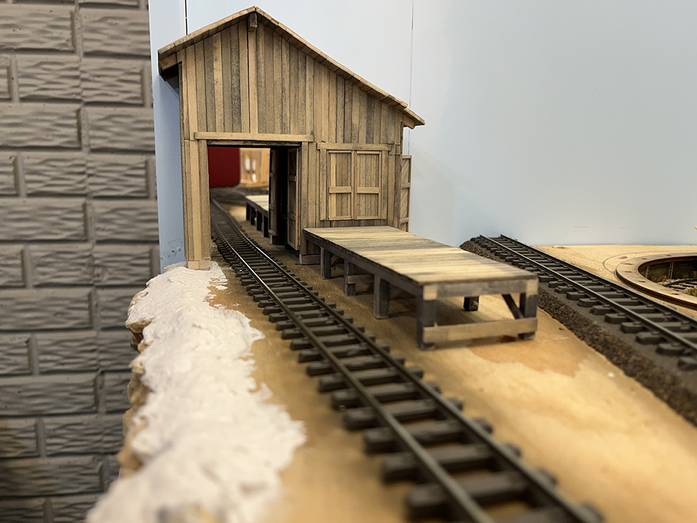 Brown building with brown railroad track running through its open door alongside a weathered wood loading dock.