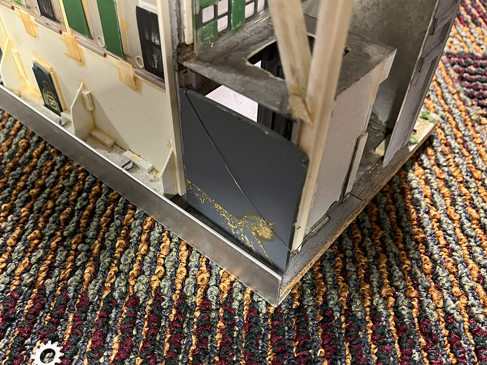 Add depth to structures using mirrors: Exposed back of white plastic model building with green doors and windows and a large gray square held with silver wire.