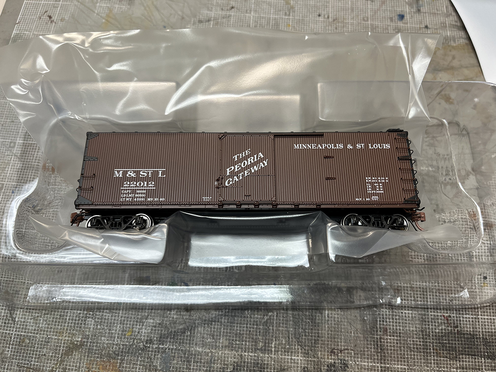 Car Swap Project Part 2: Stripping lettering from models: Brown model boxcar in clear plastic package resting on gray workbench