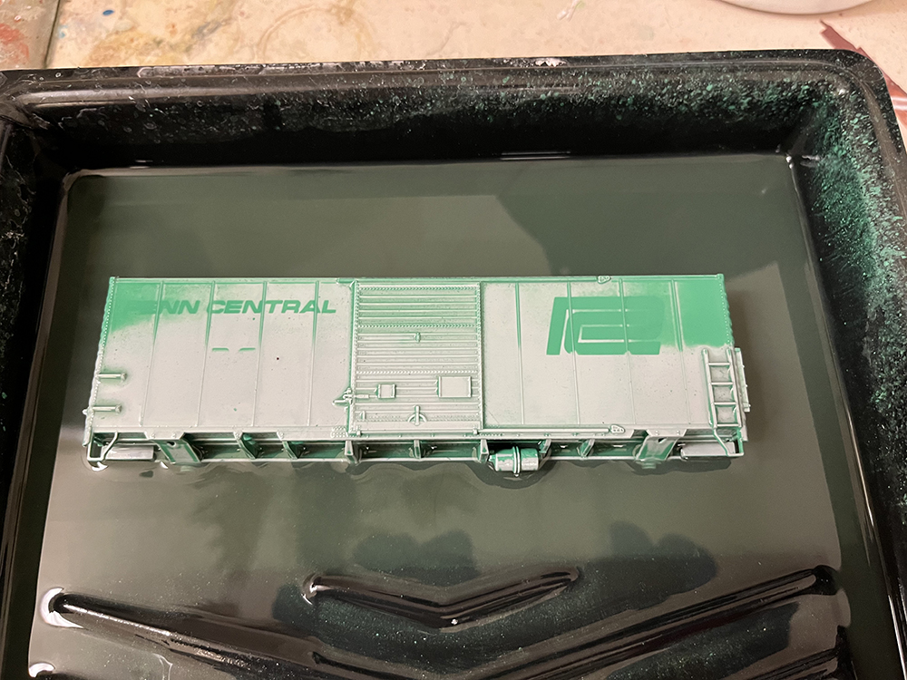 Gray and green plastic boxcar shell soaking in liquid in black paint tray
