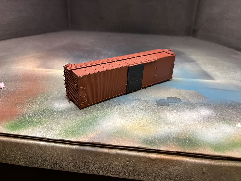 Reddish-brown plastic boxcar with black door resting in grimy spray-painting booth
