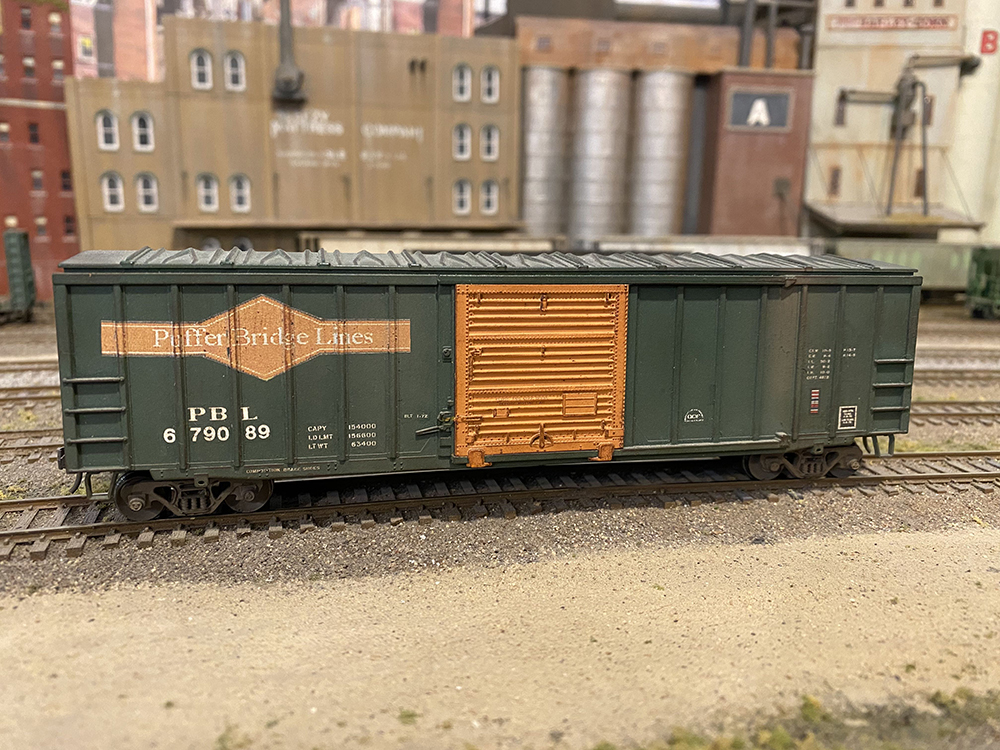 Green model boxcar with orange door and white lettering on finished model railroad track.