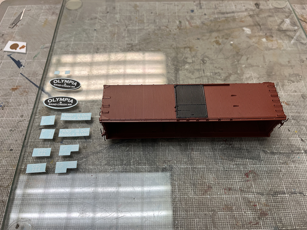 Car Swap part 8 – Olympia cars and liquid rust techniques: Small paper decals and plain brown boxcar model on a piece of clear glass.