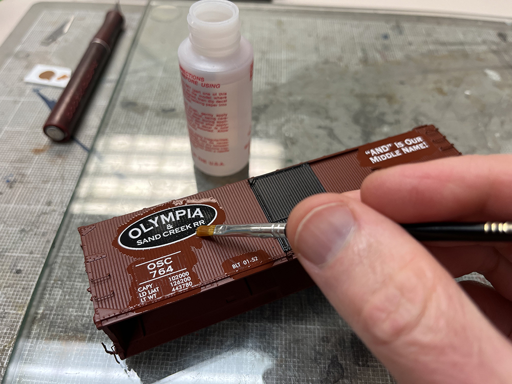 Car Swap part 8 – Olympia cars and liquid rust techniques: Paintbrush applying liquid to small decals and plain brown boxcar model