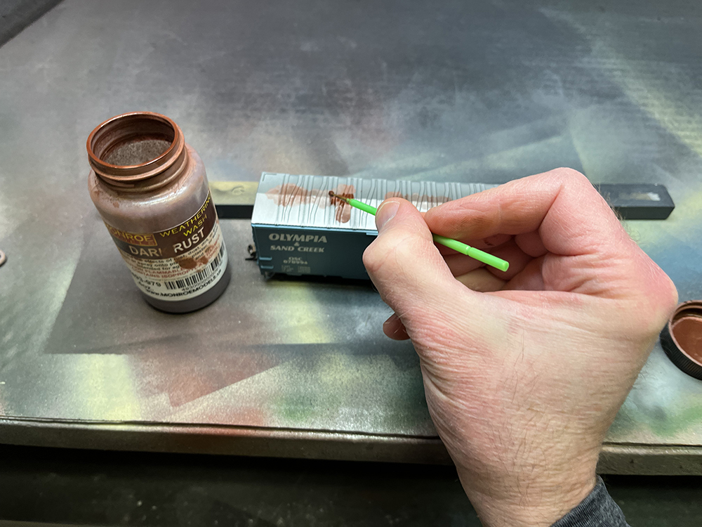 Hand holding green paint brush applying rust solution to model boxcar