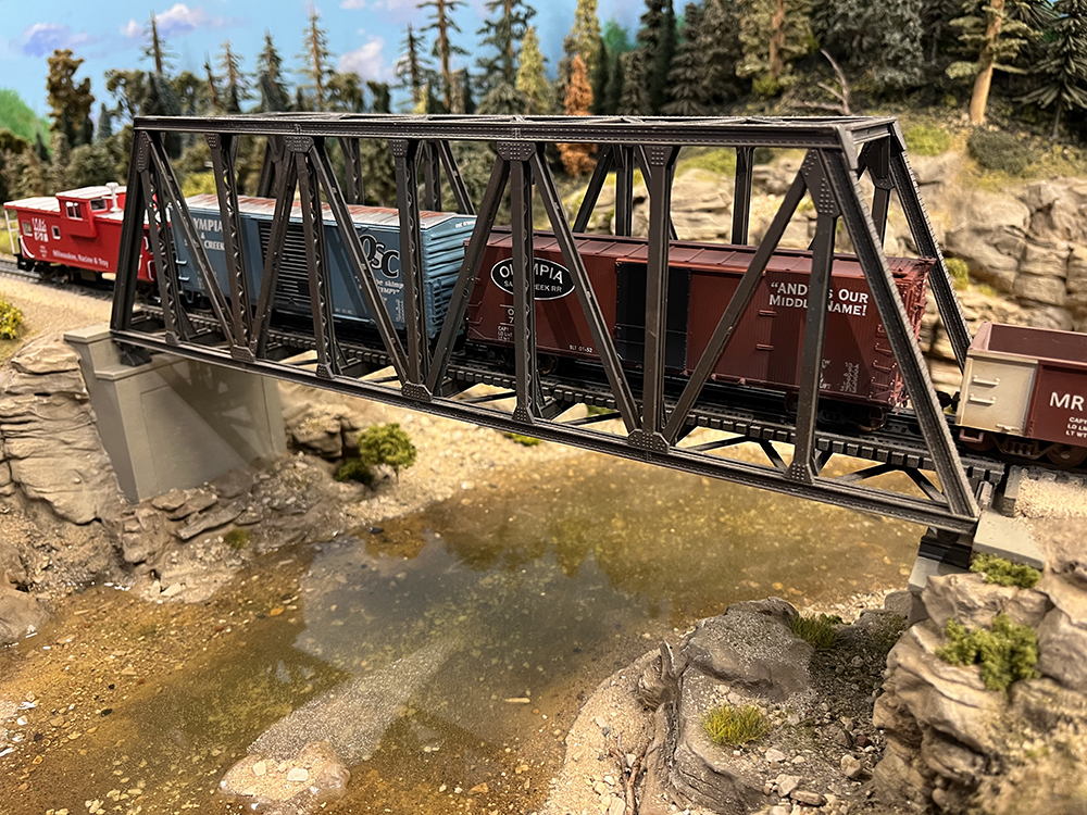 Car Swap Project part 9: Lessons learned: Red caboose and brown and blue boxcars on truss bridge over clear river on a model railroad.