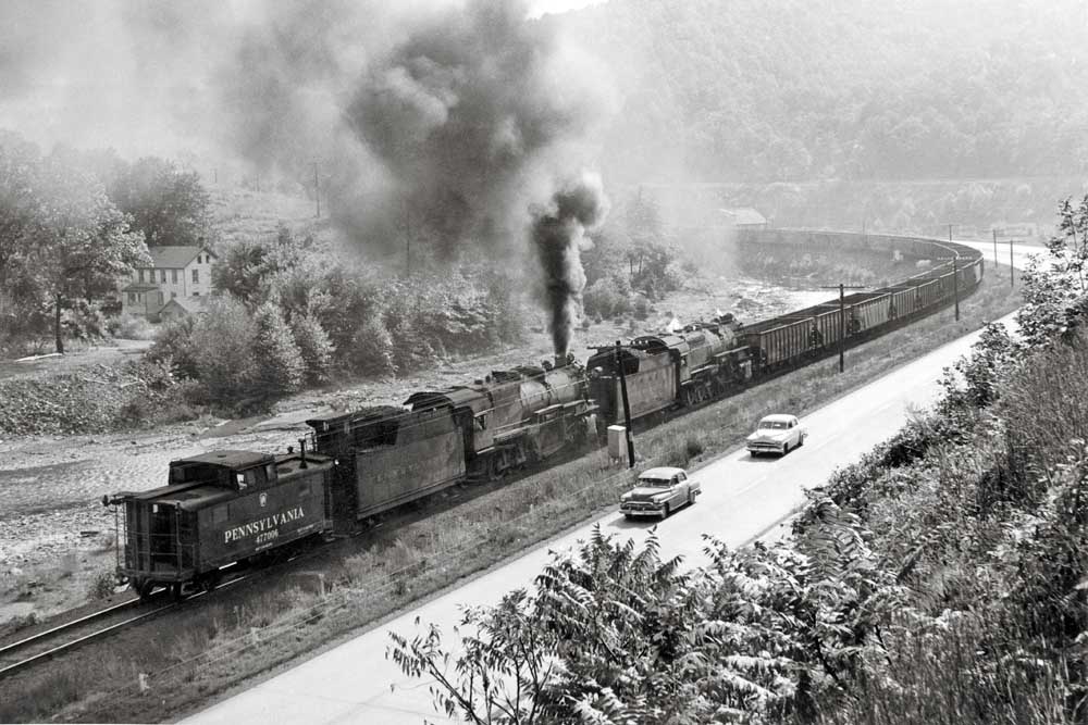 Two steam locomotives shove on a freight train in a valley