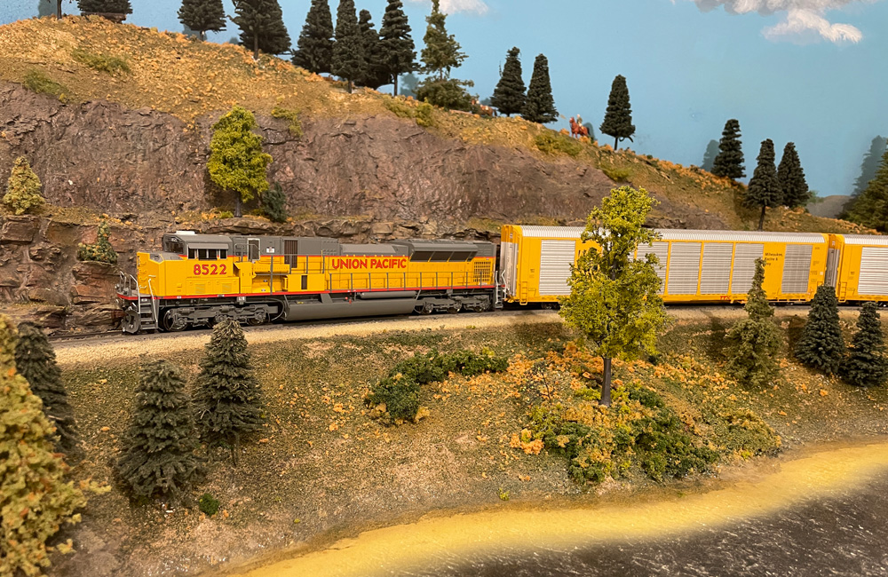 A large, modern, yellow-and-gray Union Pacific diesel rounds a curve on an HO scale layout with yellow auto rack cars in tow