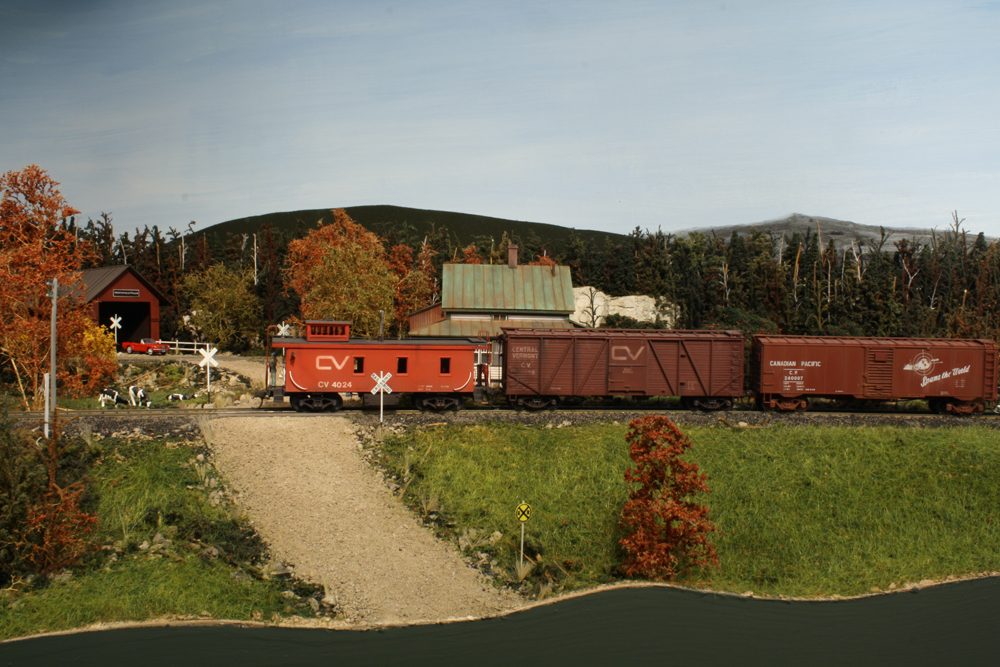 Model boxcars and a caboose pass in front of a covered bridge