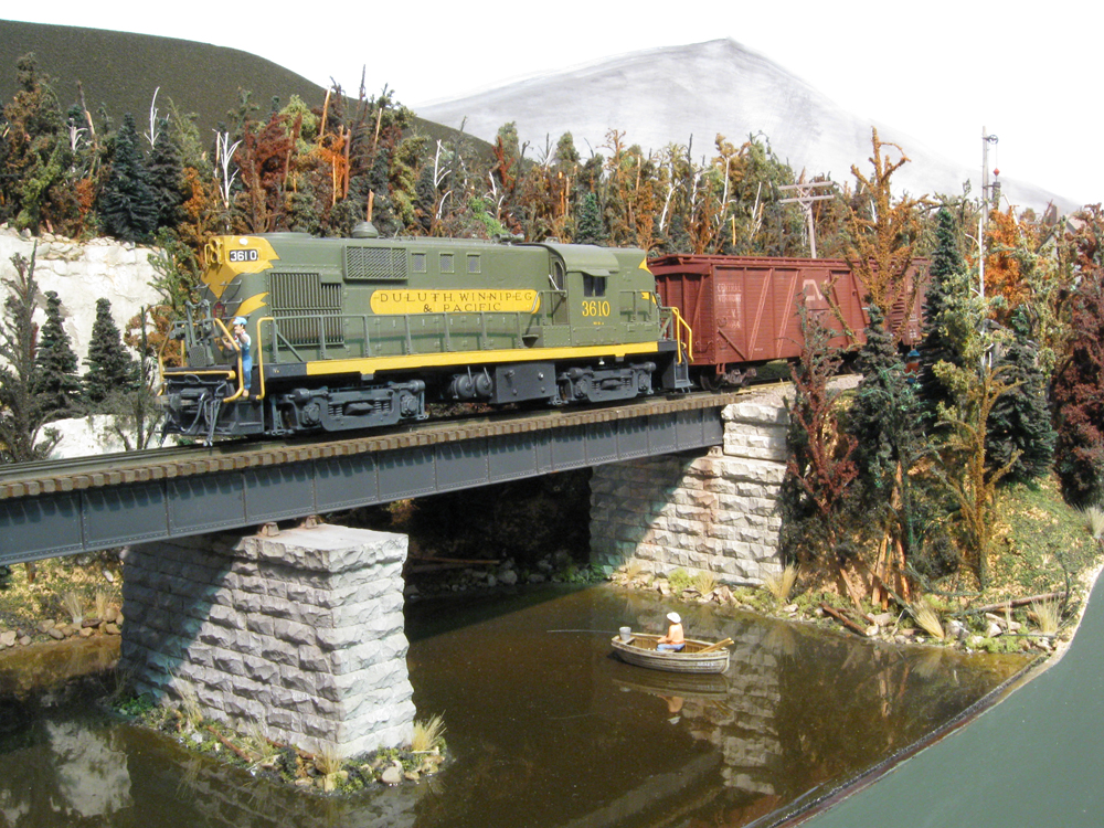 A model of a green diesel locomotive leads a train across a bridge