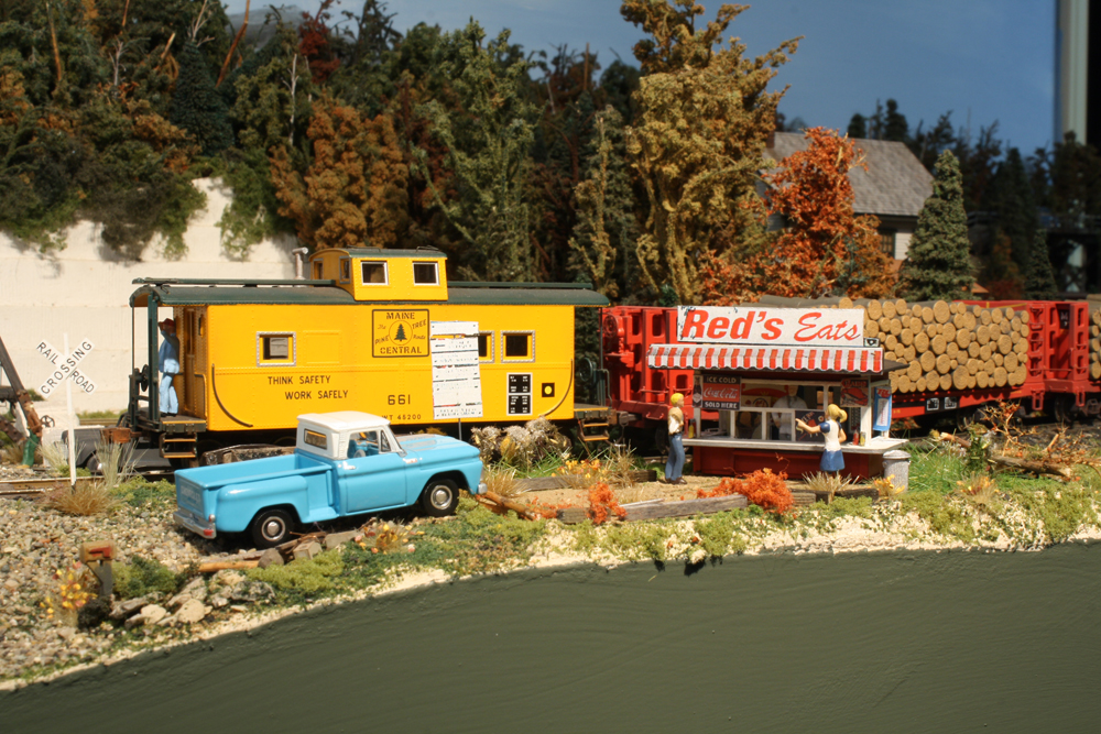 A yellow caboose passes behind a model restaurant