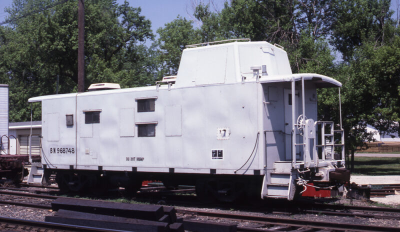 Color photo of caboose painted light gray with small windows and rooftop air conditioners.