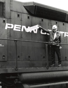 man in hardhat standing on locomotive gangway holding a sign reading "Conrail."