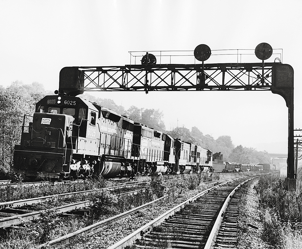 Black and white image of locomotives pulling a freight train under a signal bridge.