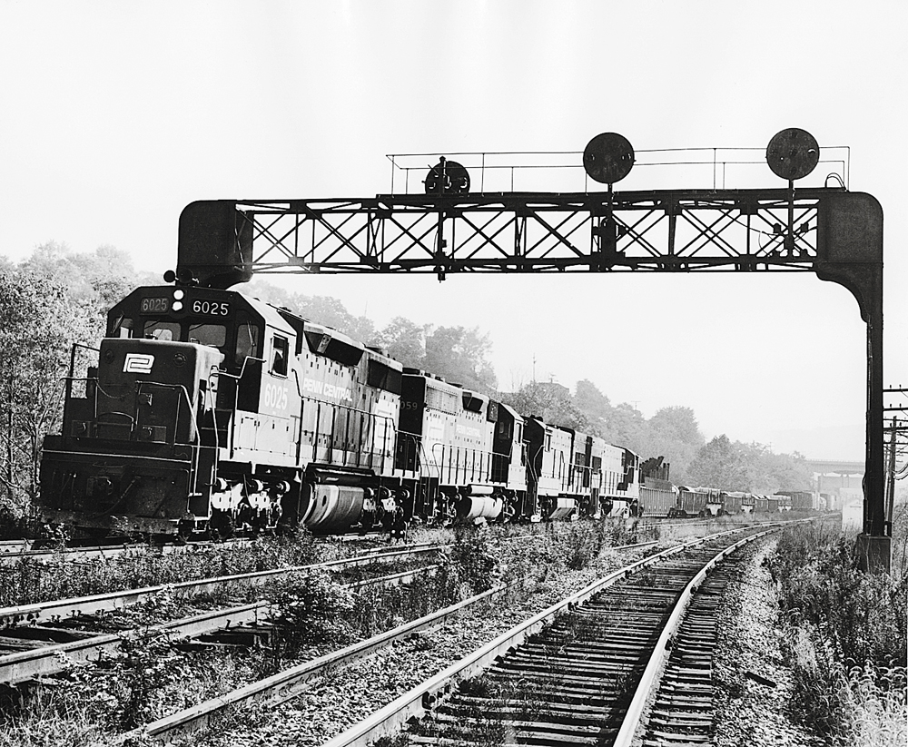Black and white picture of a freight train under a signal bridge.