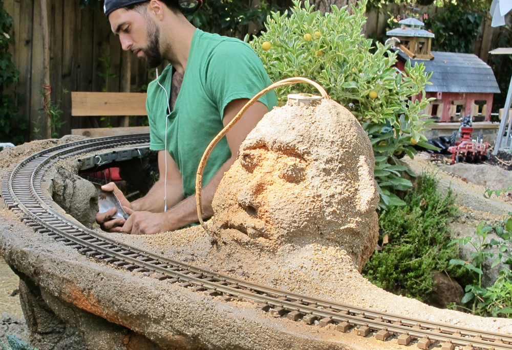 man on garden railroad near sculpture of human head