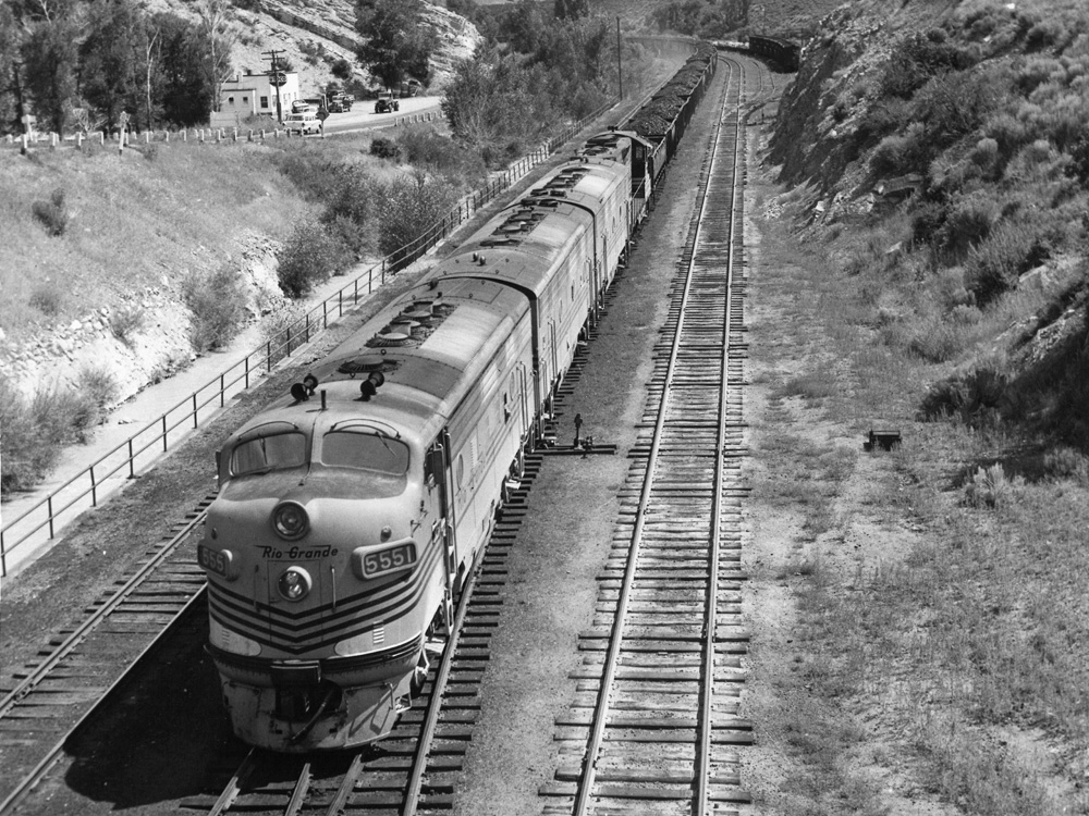A black-and-white photo of a long coal train approaching the camera, seen from a bridge over the tracks