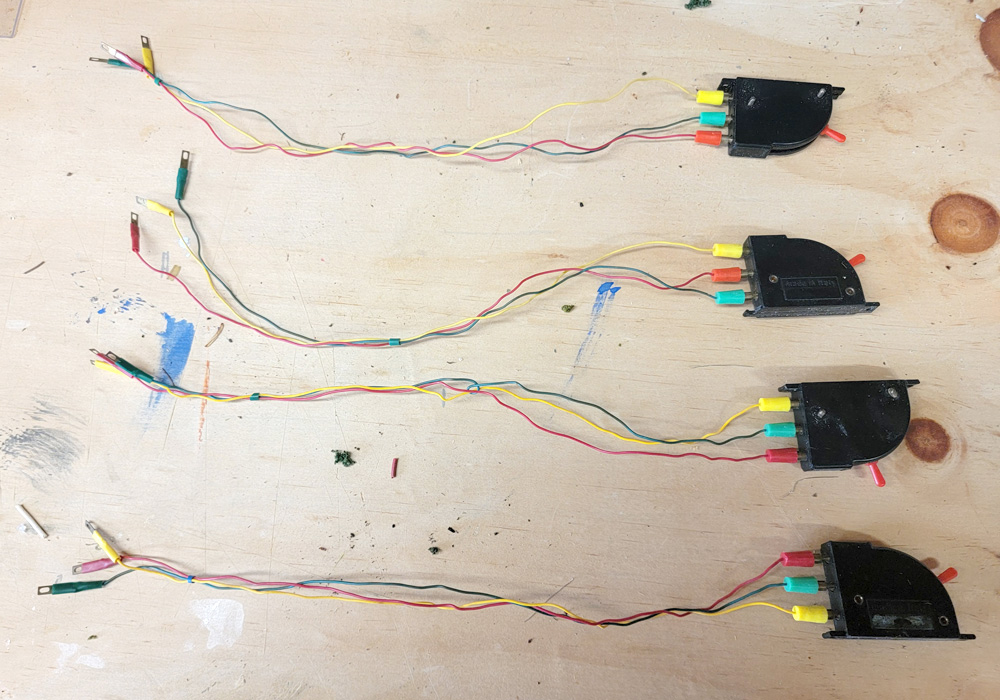 Four black rectangular devices with red lever handles and multicolored wires lie on a plywood surface