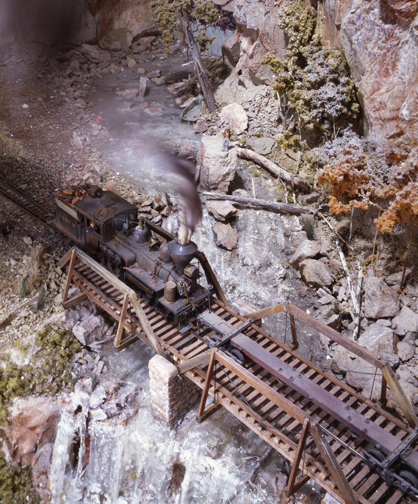 Model train on wood bridge over rushing waterfall in rugged mountain scene.
