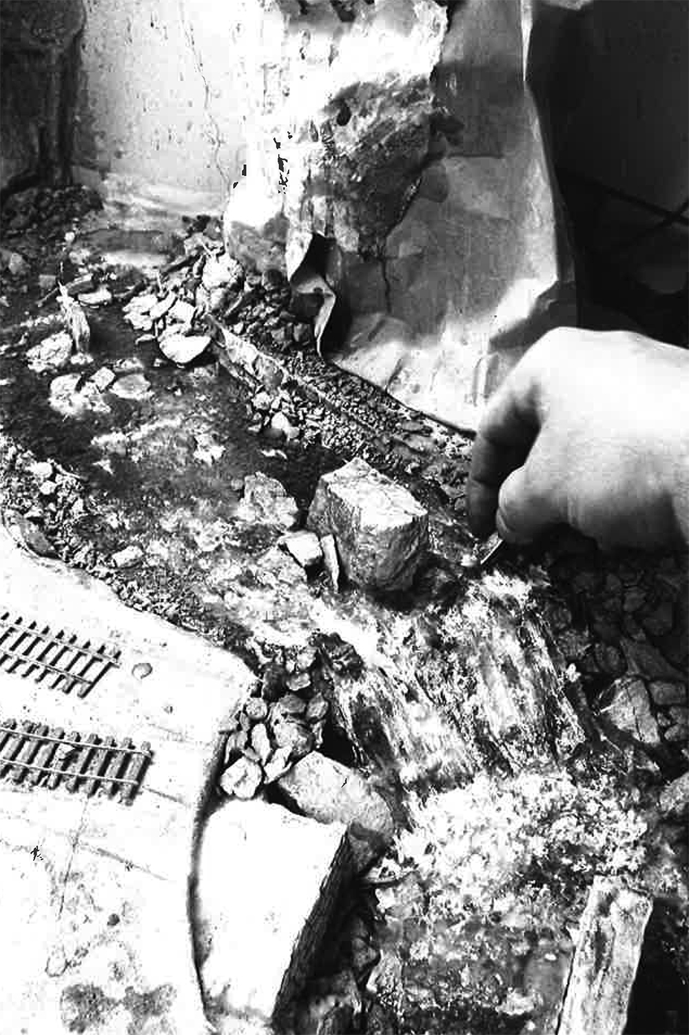 black and white photo showing hand holding ice pick and using it to tease small waves into curing resin water on a model railroad stream