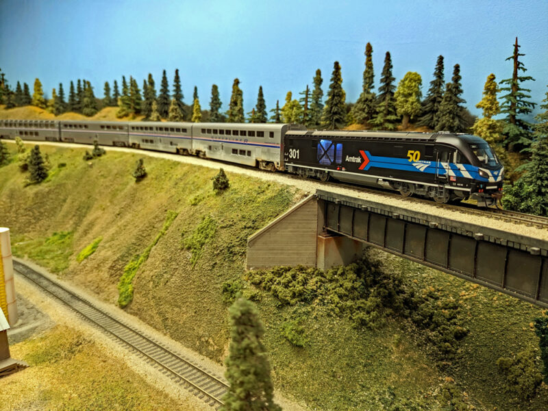 A black model diesel locomotive pulls a modern passenger train down a grade, showing why freight locomotives can’t always pull modern passenger trains.