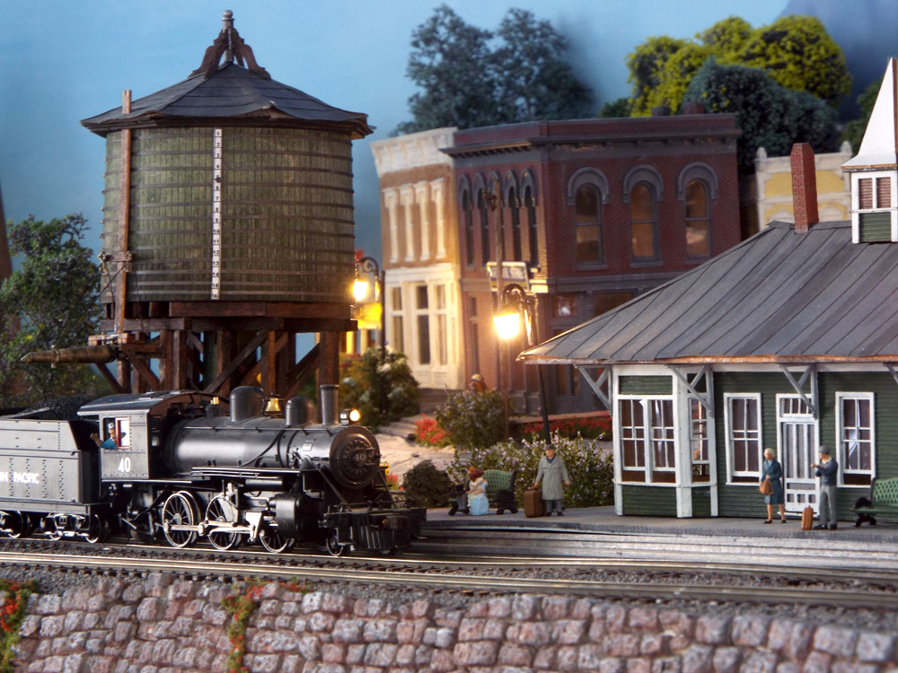 At twilight, a black steam engine refills its tender from the water tank in front of a green and white depot