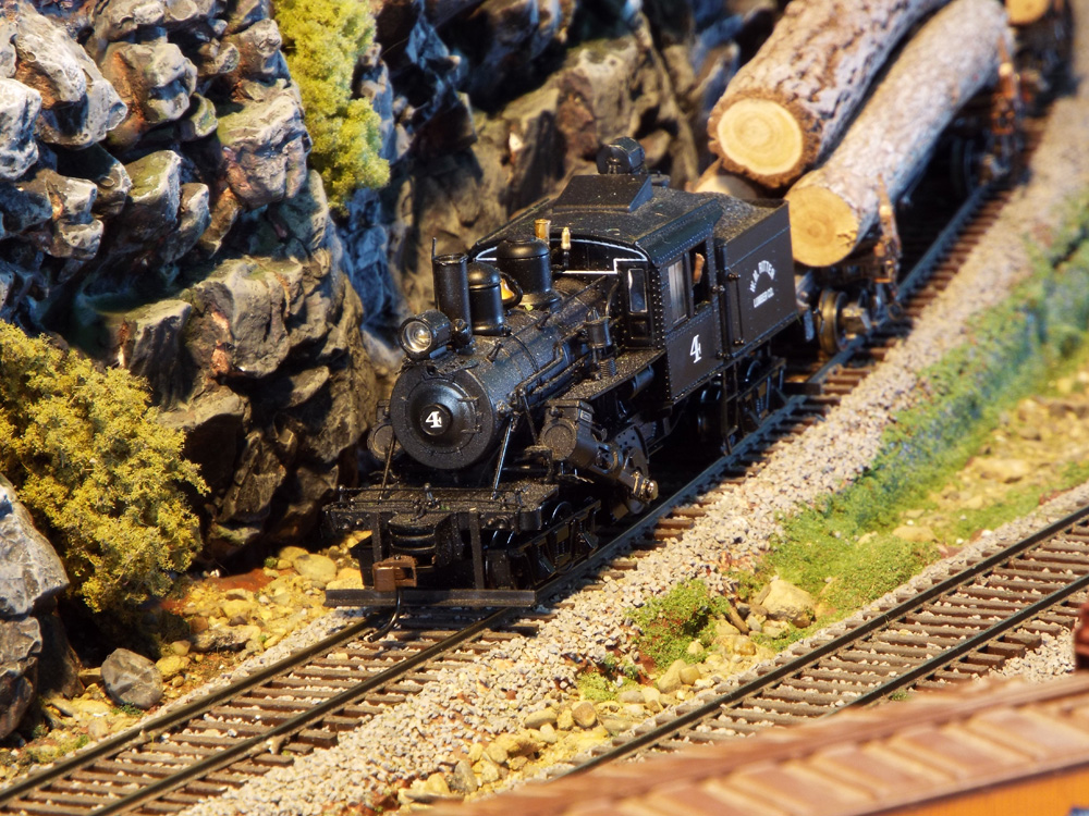 A black geared steam engine leading a log train rolls downgrade past a rock cut