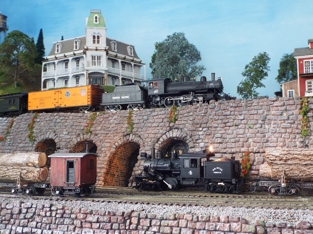 A log train arrives on a lower track while on a retaining wall behind it a mixed train rolls in the other direction