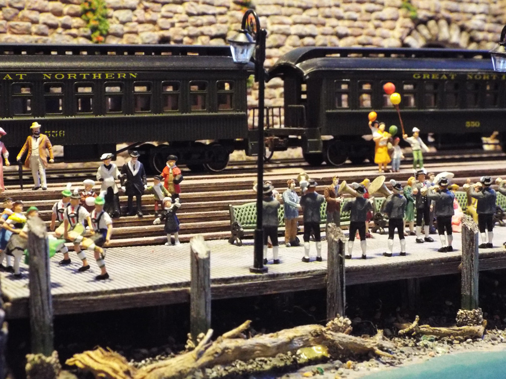 A festive crowd listens to a brass band on the town dock while passenger cars wait behind them