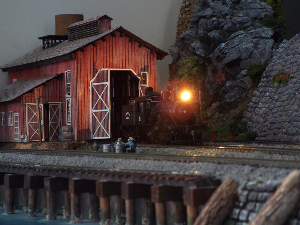 At twilight, a steam locomotive’s headlight shines brightly as it emerges from a red wood enginehouse